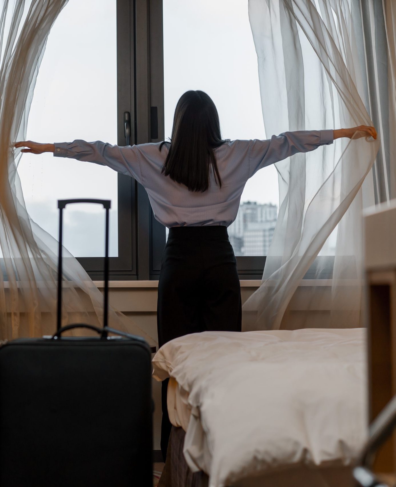 young woman opening curtain while standing luxury hotel room looking through window enjoying city skyscrapers close up