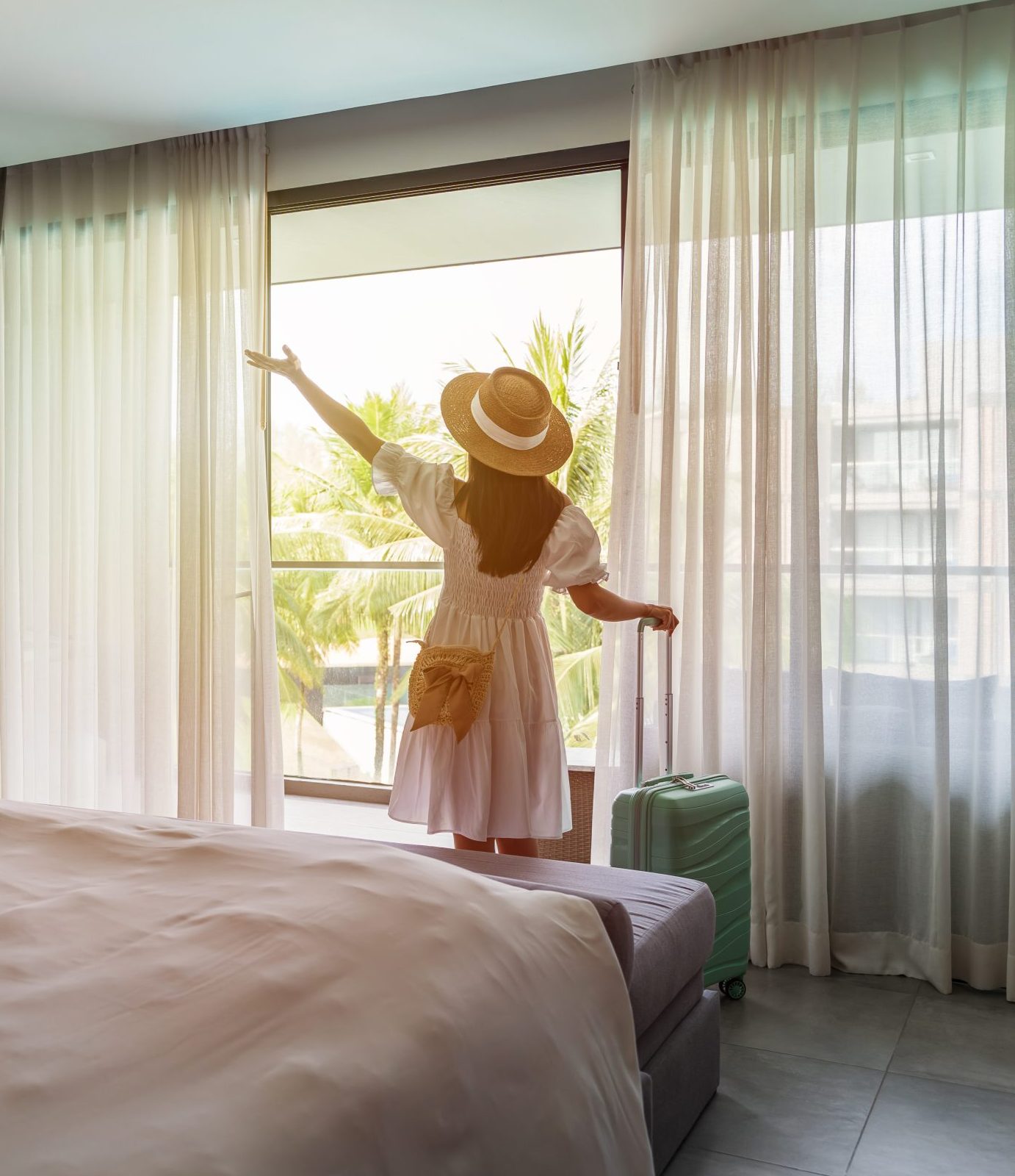 Young woman traveler opening the curtains and looking at the view from the window of a hotel room while on summer vacation, Travel lifestyle concept